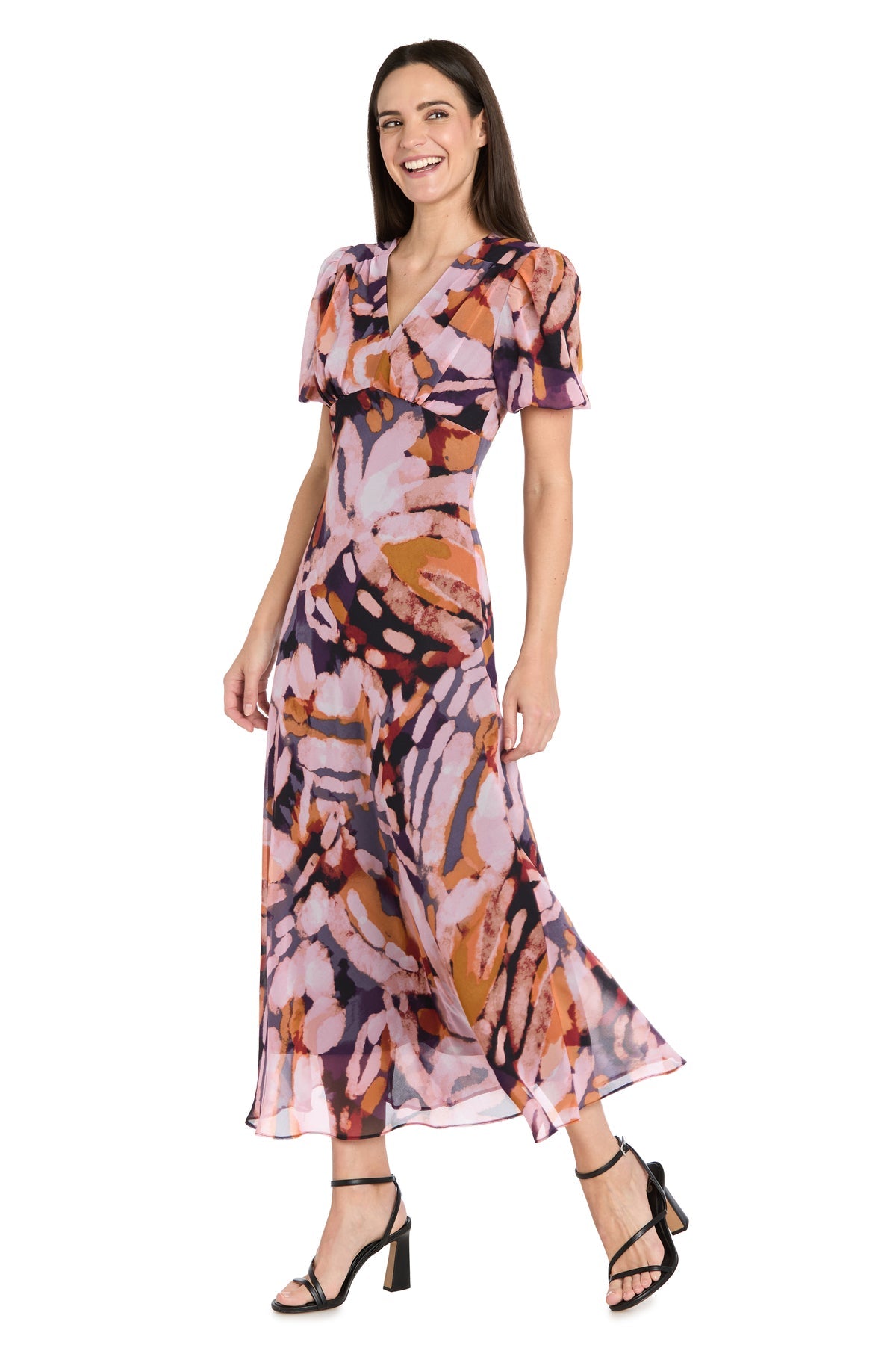 A woman models the R&M Richards 4161 Short Sleeve Print Party Dress, featuring an abstract multicolor pattern, for a semi-formal or wedding guest look. She pairs it with black heeled sandals and smiles against a white background.